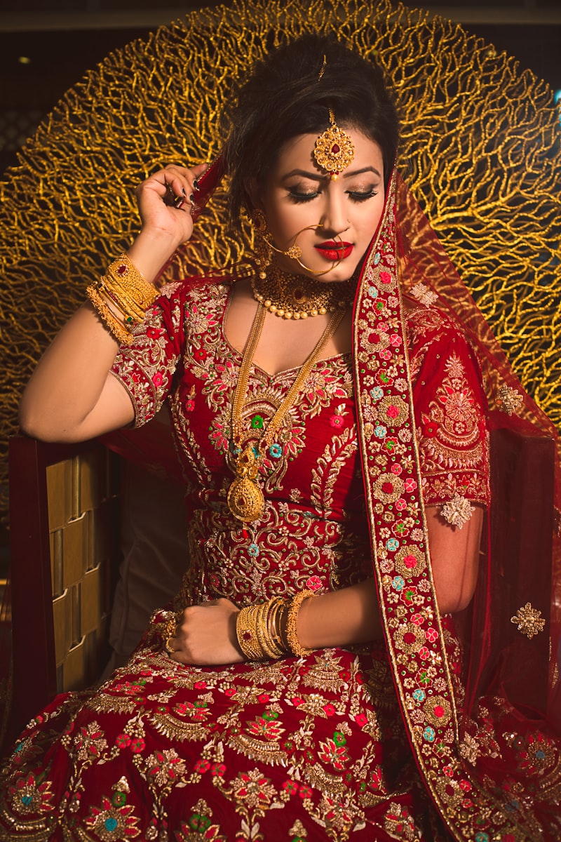 Bride in embroidered red and gold attire with completed bridal makeup, seated before a gold backdrop
